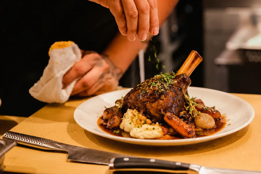 A chef seasoning a braised lamb shank at the pass